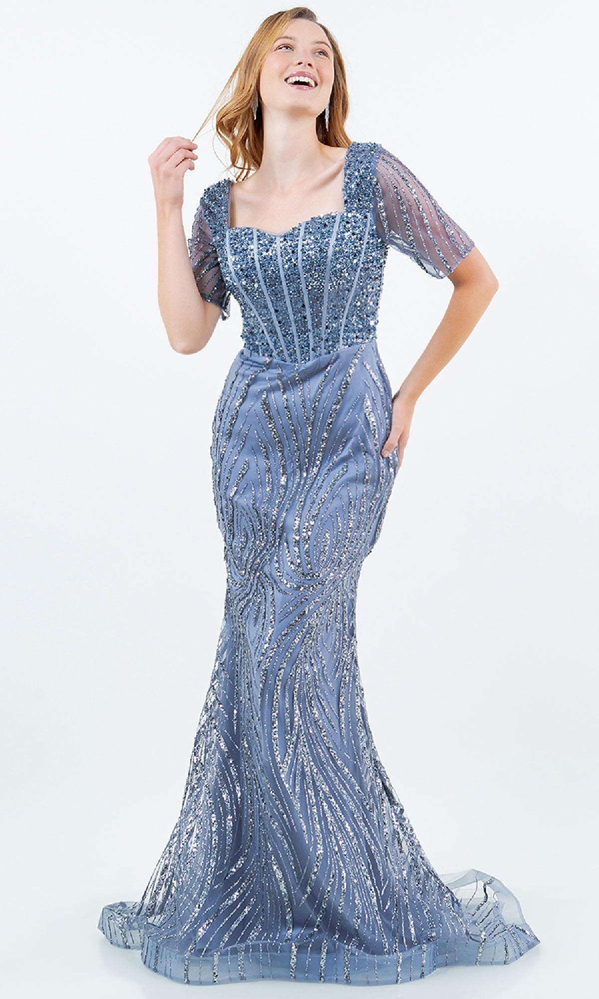 A woman wears the Long Sequin Formal Gown: Cinderella Couture 8293M—a blue, floor-length mermaid dress with intricate beadwork, short sheer sleeves, and a square neckline—standing against a plain background.